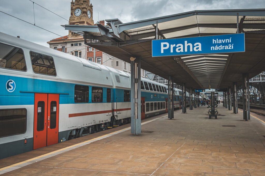 Dvoupodlažní elektrické jednotky CityElefant Českých drah v modrobílém nátěru stojící u nástupiště pražského hlavního nádraží. V popředí je výrazná modrá nádražní cedule s názvem stanice.