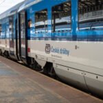 Souprava osobních vozů Českých drah v modrobílém nátěru stojící u nástupiště železniční stanice. Na vozech je viditelné logo národního dopravce