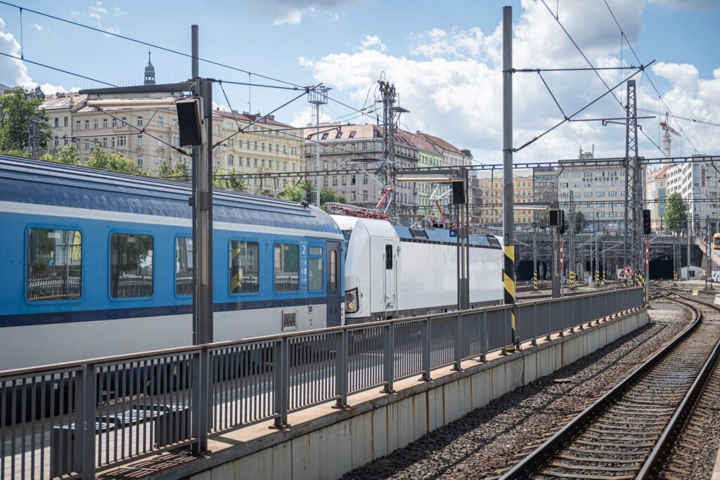 Souprava Českých drah s modrobílými osobními vozy a elektrickou lokomotivou ve stanici Praha hlavní nádraží. V pozadí jsou železniční koleje, trakční vedení a městská zástavba