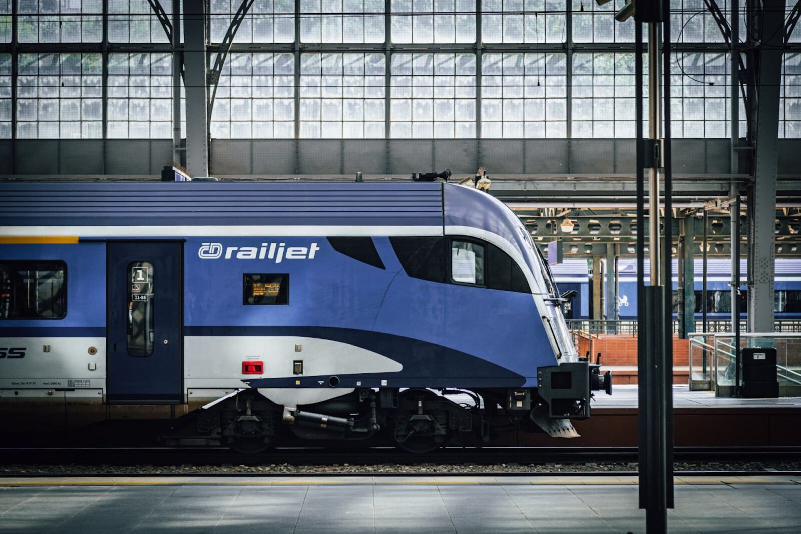 Detail řídicího vozu soupravy railjet Českých drah v modrobílém nátěru na pražském hlavním nádraží, pod historickou prosklenou nádražní halou