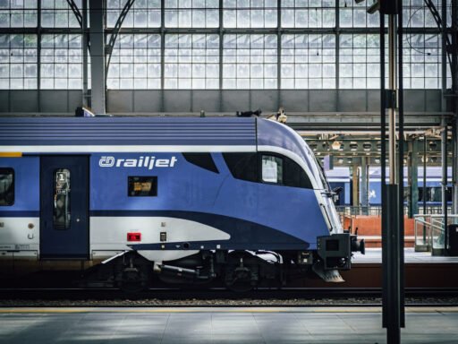 Detail řídicího vozu soupravy railjet Českých drah v modrobílém nátěru na pražském hlavním nádraží, pod historickou prosklenou nádražní halou