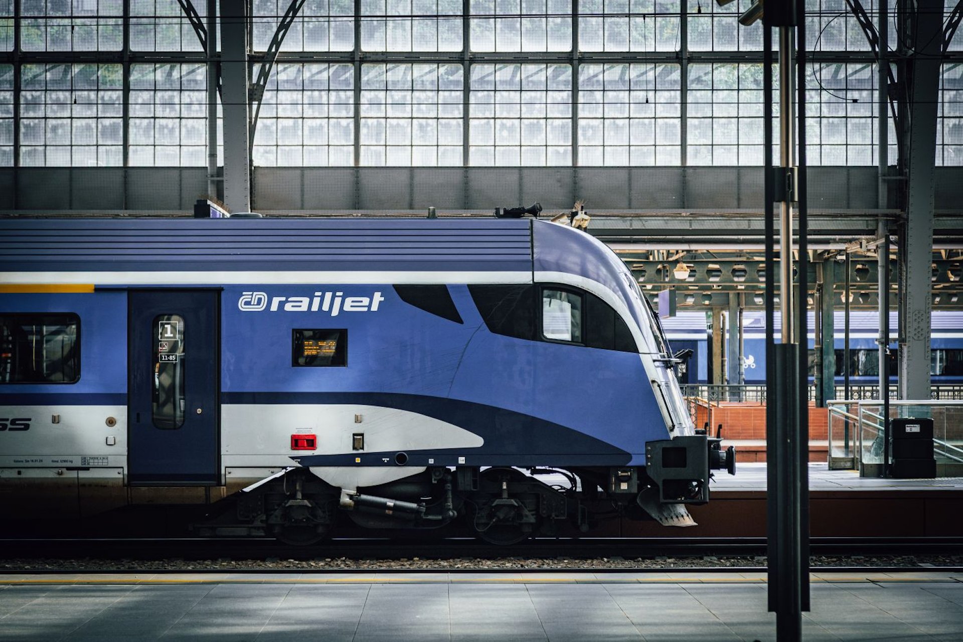 Detail řídicího vozu soupravy railjet Českých drah v modrobílém nátěru na pražském hlavním nádraží, pod historickou prosklenou nádražní halou