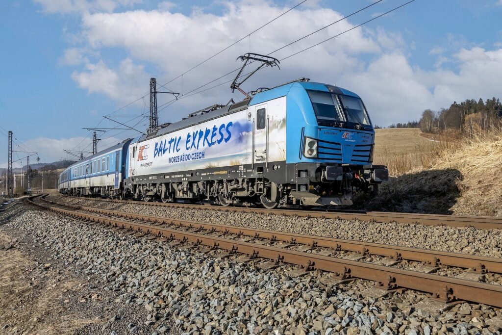 Mezinárodní vlak Baltic Express vedený elektrickou lokomotivou Siemens Vectron v modrobílém nátěru PKP Intercity projíždí po elektrifikované železniční trati v otevřené krajině