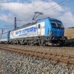 Mezinárodní vlak Baltic Express vedený elektrickou lokomotivou Siemens Vectron v modrobílém nátěru PKP Intercity projíždí po elektrifikované železniční trati v otevřené krajině