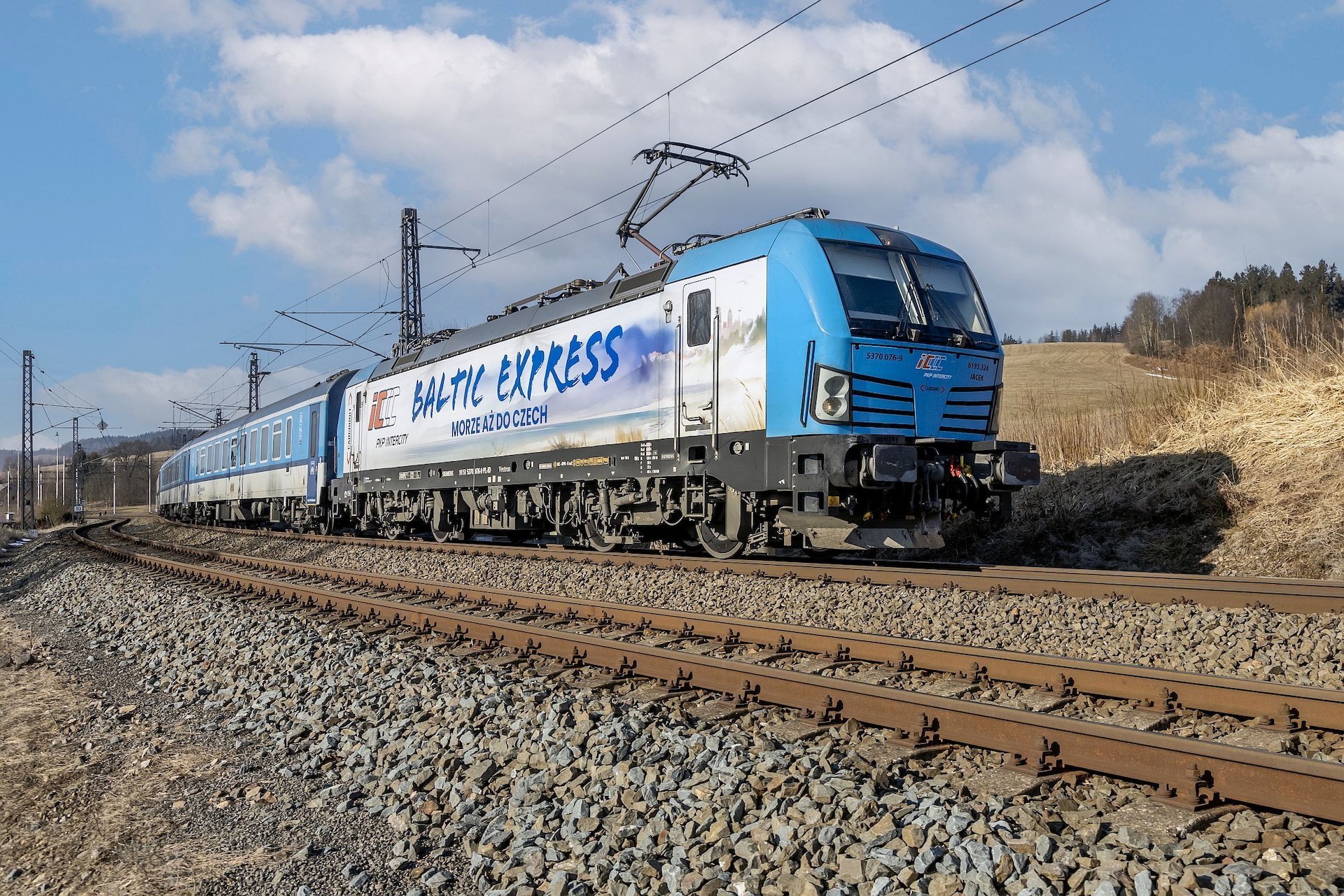 Mezinárodní vlak Baltic Express vedený elektrickou lokomotivou Siemens Vectron v modrobílém nátěru PKP Intercity projíždí po elektrifikované železniční trati v otevřené krajině