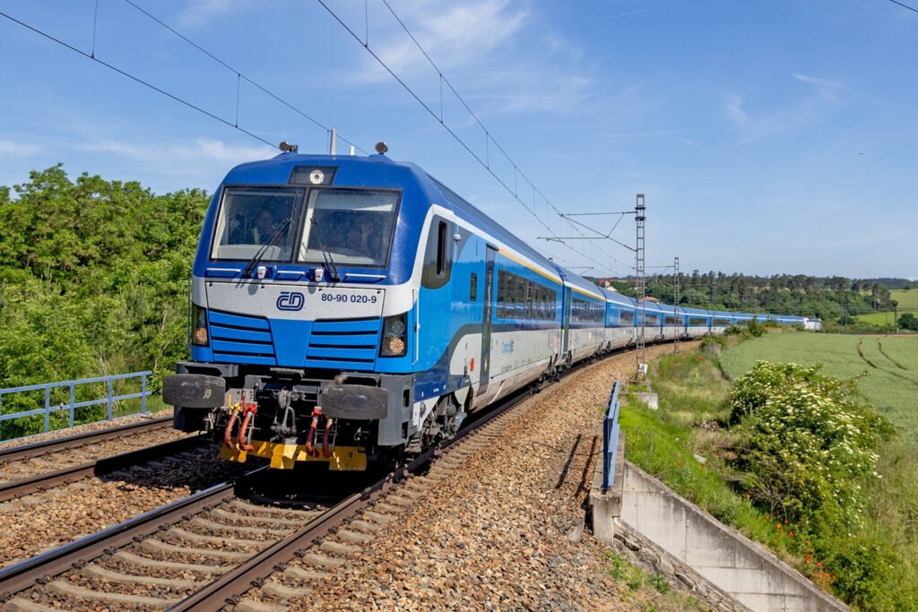 Moderní souprava ComfortJet Českých drah v modrobílém nátěru projíždí obloukem elektrifikované železniční trati v zelené krajině