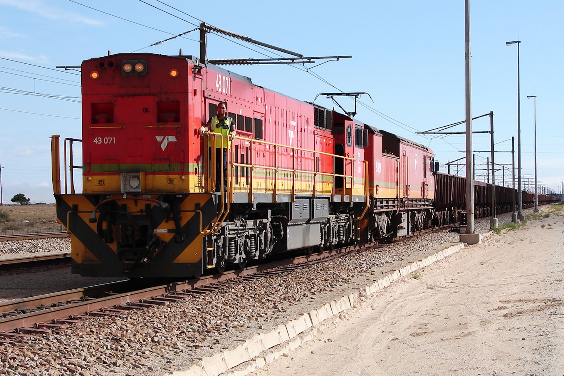 Dieselelektrická lokomotiva řady 43-000 dopravce Transnet Freight Rail v červeném nátěru vede dlouhou soupravu nákladních vozů na železniční trati u jihoafrického města Saldanha
